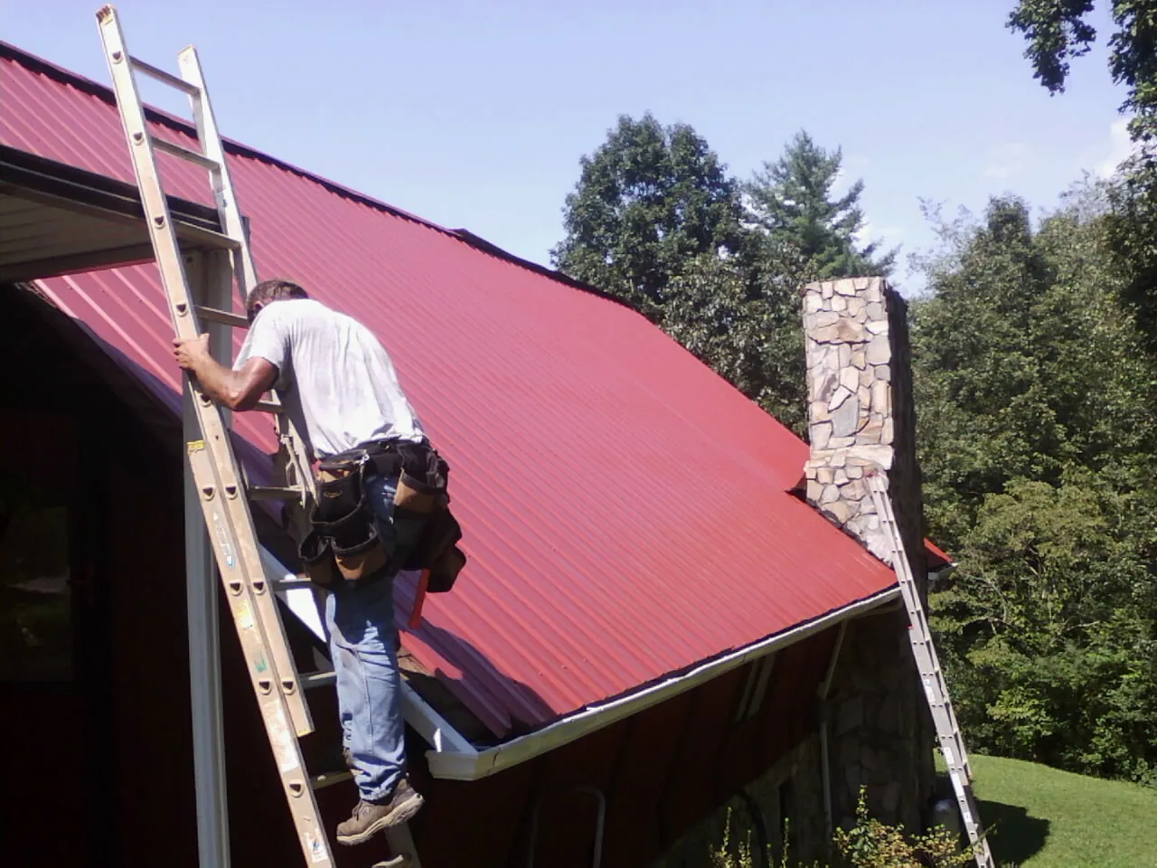 Roofer on a ladder beside a red metal roof and stone chimney — Soffit & Fascia Repair by Roofing Brothers in Scottsburg